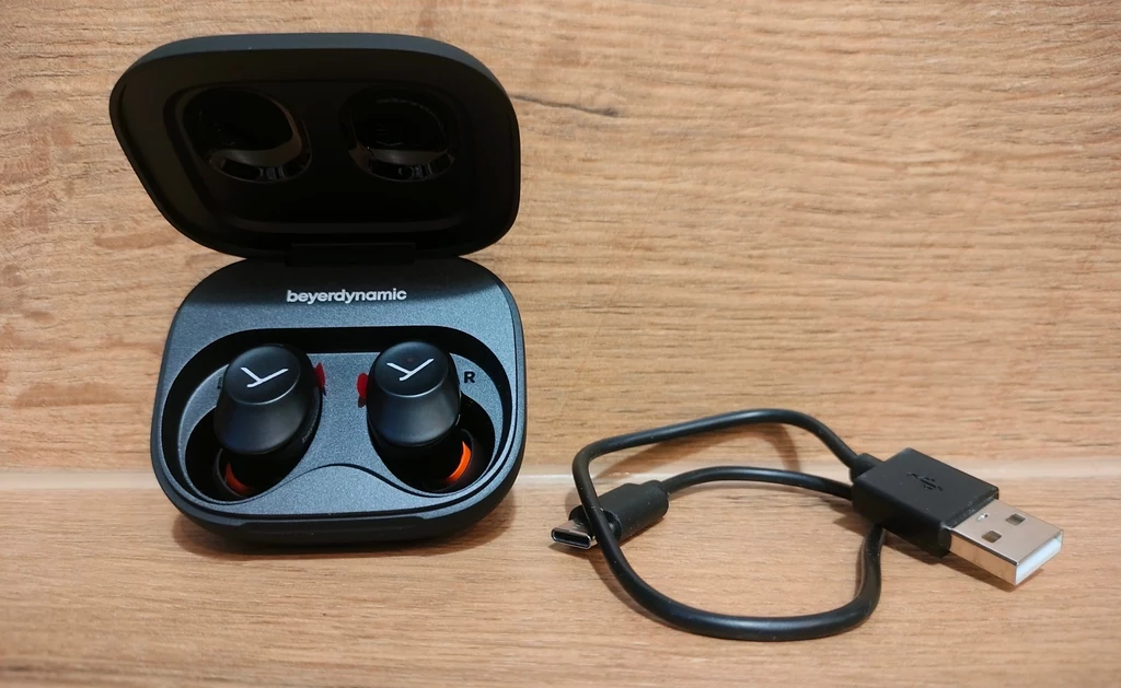 Beyerdynamic Amiron 300 Bezprzewodowe słuchawki douszne w etui ładującym marki Beyerdynamic obok czarnego kabla USB do ładowania, całość ustawiona na drewnianej powierzchni.