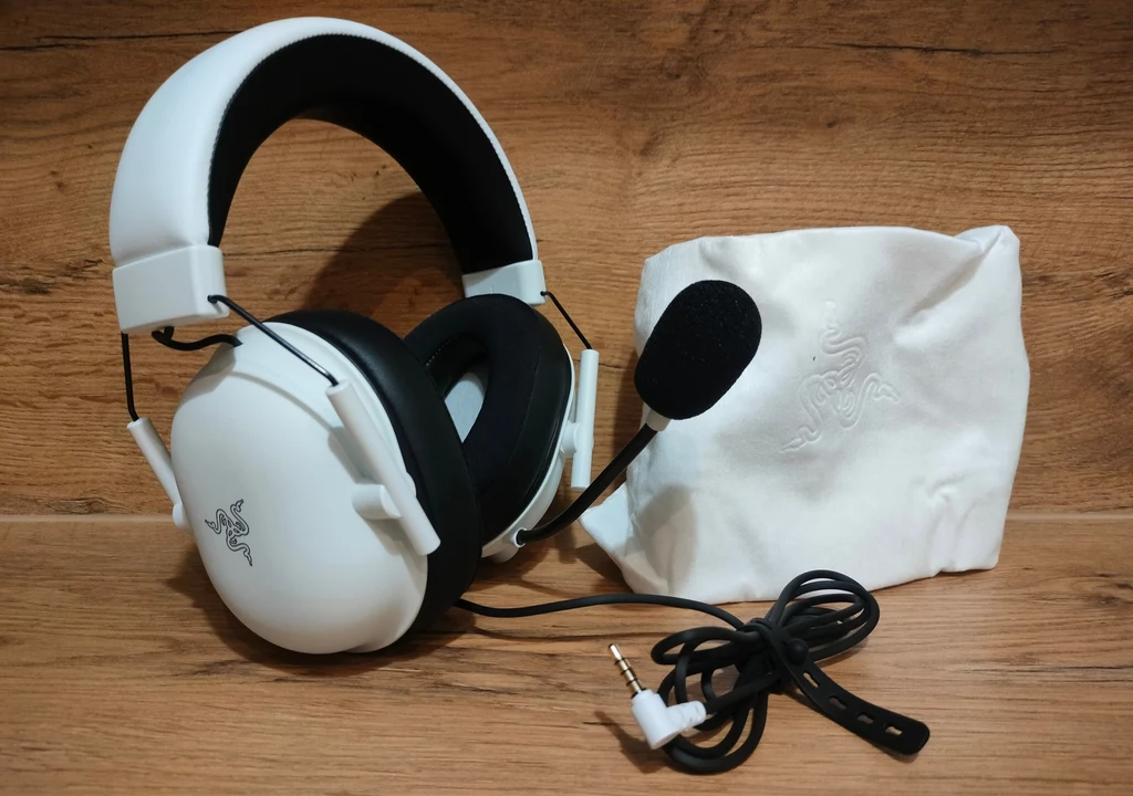 Razer BlackShark V2 X White Edition Białe słuchawki nauszne z mikrofonem na pałąku, przewodem oraz miękkim, białym etui, ustawione na tle drewnianej powierzchni.