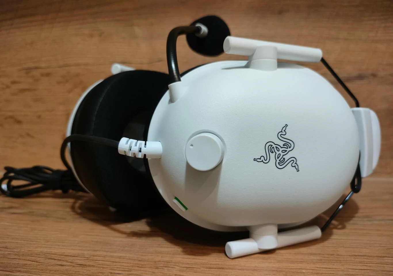 Białe słuchawki z mikrofonem marki Razer leżą na drewnianej powierzchni, widoczny jest charakterystyczny logotyp firmy oraz przewód podłączony do urządzenia.