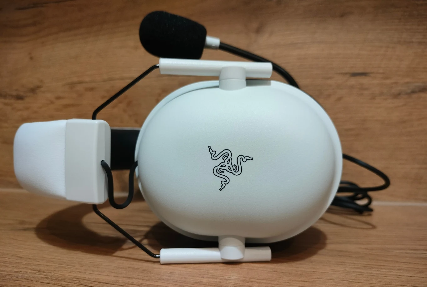 Białe słuchawki z mikrofonem nauszny, z wyraźnie eksponowanym logo marki Razer, ułożone na drewnianej powierzchni.