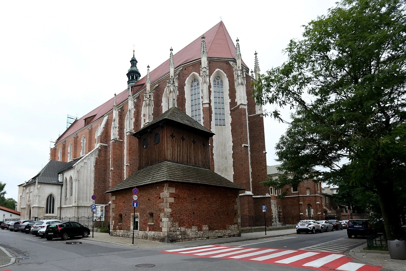 Kościół z czerwonej cegły w stylu gotyckim, z drewnianą basztą połączoną z murem, usytuowany przy ulicy z zaparkowanymi samochodami, po lewej widoczne przejście dla pieszych, otoczenie zieleni i drzew.