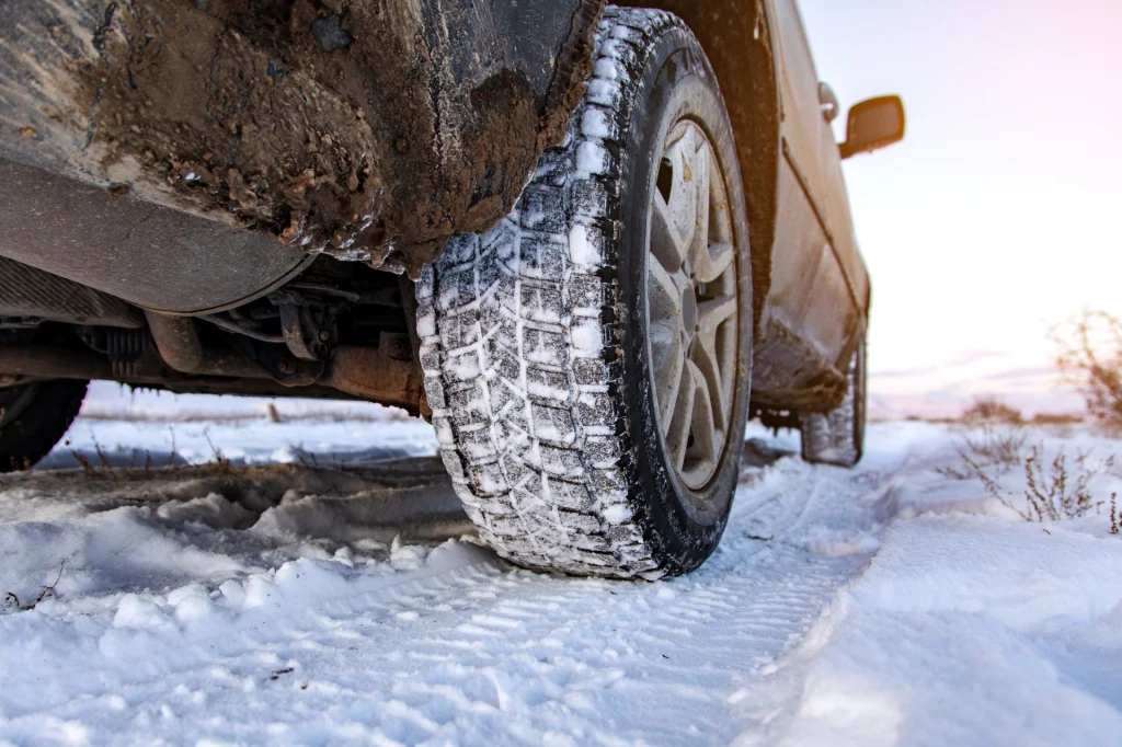 Samochód terenowy na zaśnieżonej drodze, widoczny wyraźnie bieżnik zimowej opony pokrytej śniegiem; błoto i śnieg na nadkolu pojazdu, krajobraz zimowy w tle.