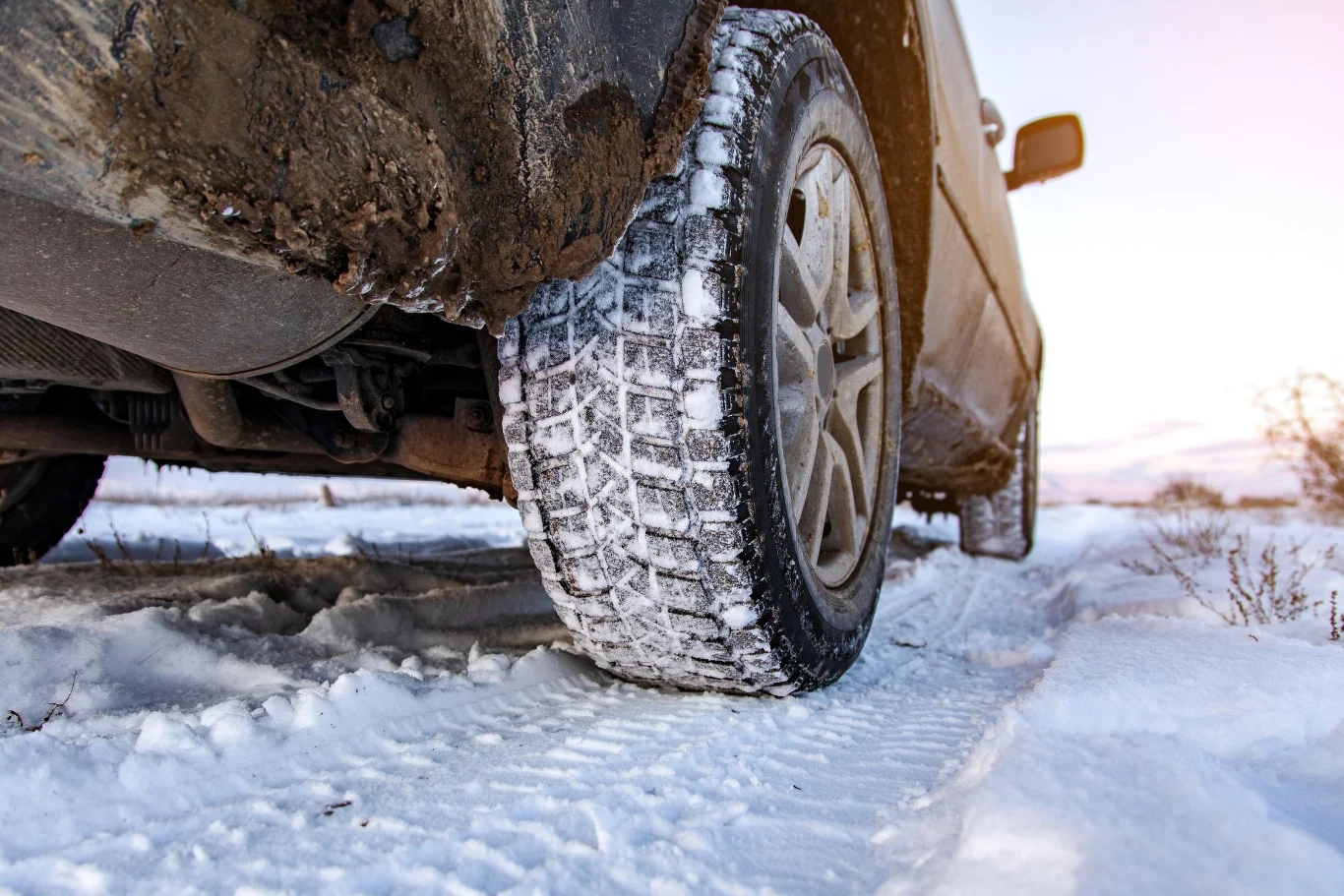 Część kierowców nawet nie wie, że da się założyć opony odwrotnie Samochód terenowy na zaśnieżonej drodze, widoczny wyraźnie bieżnik zimowej opony pokrytej śniegiem; błoto i śnieg na nadkolu pojazdu, krajobraz zimowy w tle.