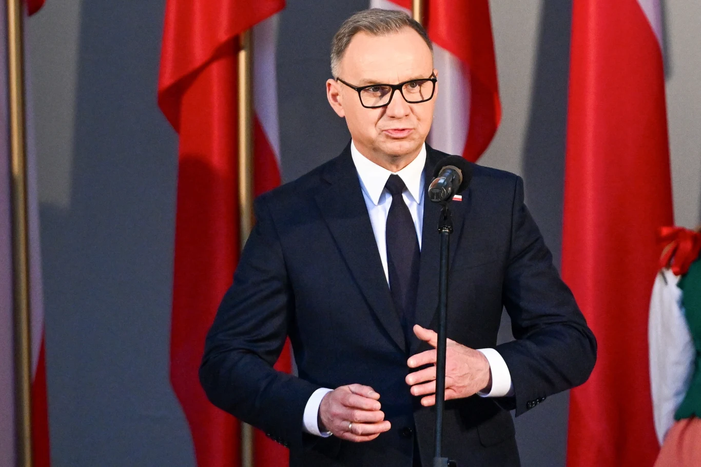 Prezydent Andrzej Duda Mężczyzna w garniturze i okularach przemawia przy mikrofonie, w tle widoczne liczne polskie flagi.