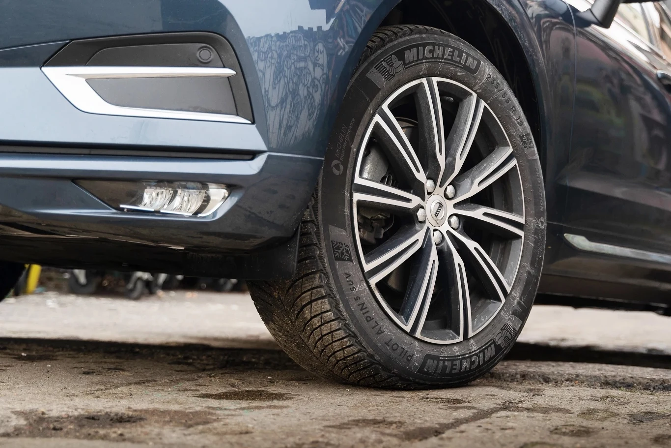 Czy za jazdę latem na zimówkach jest mandat? Zbliżenie na tylne koło pojazdu osobowego z aluminiową felgą i oponą Michelin, fragment karoserii w kolorze niebieskim, auto stoi na nierównej nawierzchni.