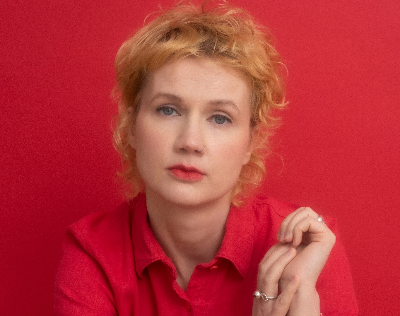 Dorota Groyecka Portret kobiety o jasnej karnacji z blond falującymi włosami, ubrana w czerwoną koszulę, pozująca na tle ściany w tym samym kolorze, z delikatnym makijażem i spokojnym wyrazem twarzy.