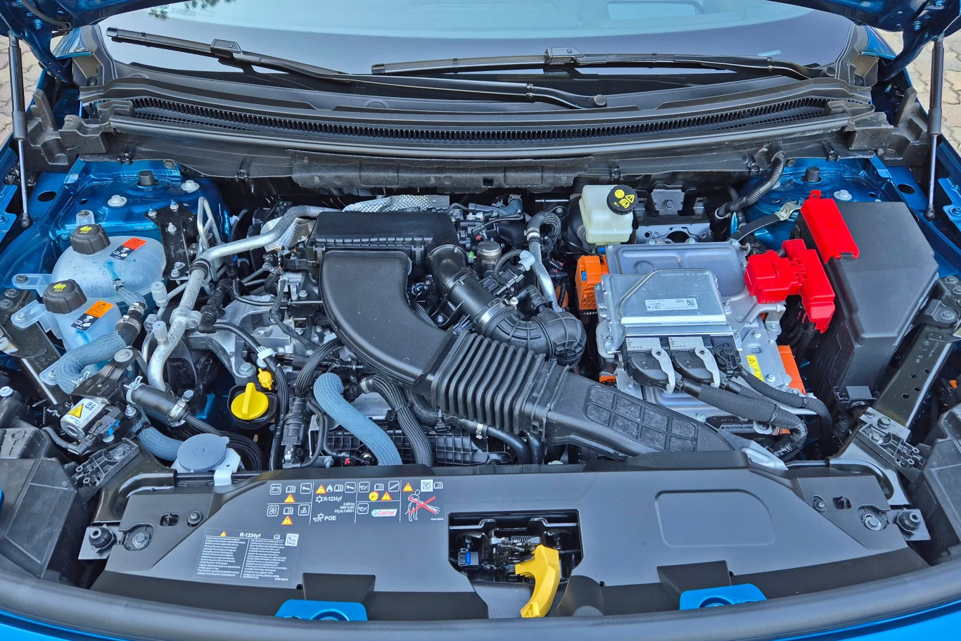 Widok nowoczesnej komory silnika samochodowego, widoczne liczne podzespoły mechaniczne i elektroniczne, elementy układu chłodzenia, przewody oraz akumulator w czerwonej obudowie.
