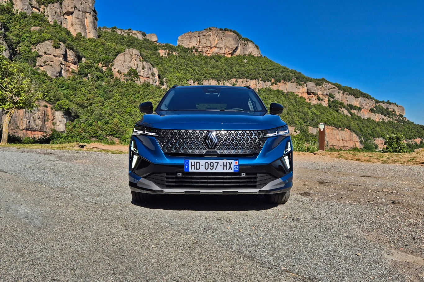 Niebieski samochód marki Renault zaparkowany na otwartym terenie żwirowym, w tle zielone wzgórza i formacje skalne pod czystym, niebieskim niebem.