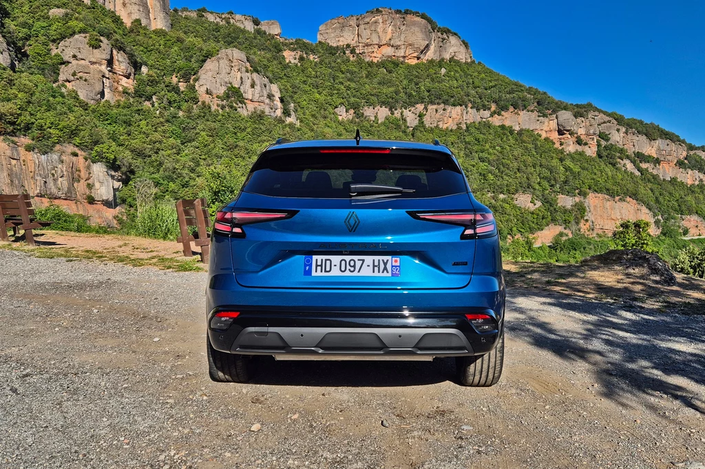 Renault Austral Niebieski samochód osobowy stojący tyłem na parkingu żwirowym, otoczony malowniczymi skałami i zielonymi wzgórzami w tle, przy słonecznej pogodzie.