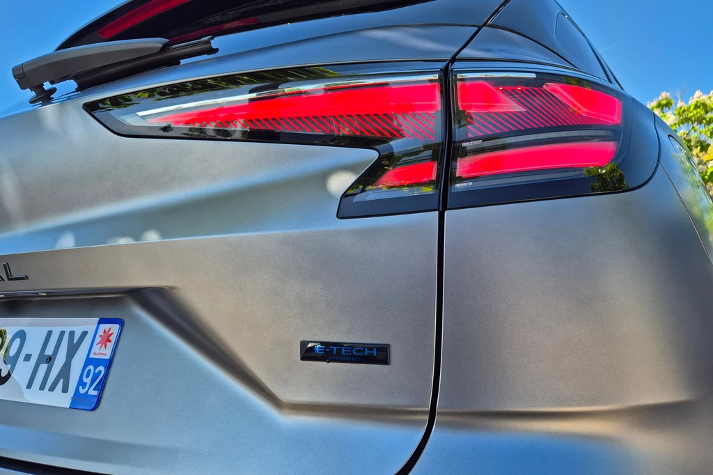 Renault Austral Nowoczesny samochód w kolorze srebrnym, widoczne tylne światła LED oraz emblemat E-Tech na klapie bagażnika, a także fragment tablicy rejestracyjnej, całość sfotografowana pod niebieskim niebem