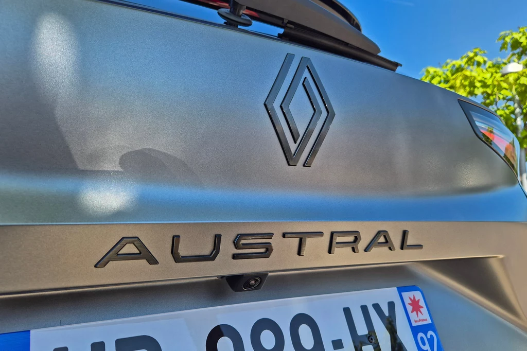 Renault Austral Logo Renault oraz napis AUSTRAL na klapie bagażnika samochodu, fragment rejestracji z niebieskim paskiem i symbolem regionu, tło z niebieskim niebem oraz zielonym liściem drzewa.