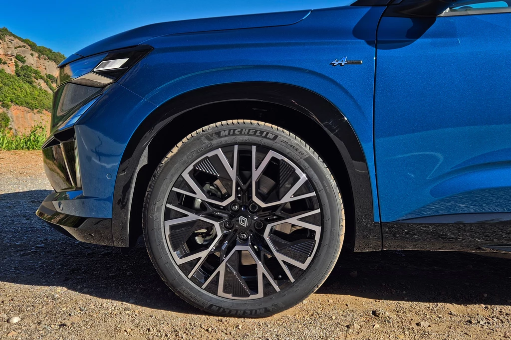 Renault Austral Przednie koło samochodu osobowego z charakterystyczną felgą aluminiową i oponą Michelin, karoseria pojazdu w kolorze niebieskim z widocznym detalem oznaczenia modelu, tło z nieostrymi elementami natury oraz drogą.