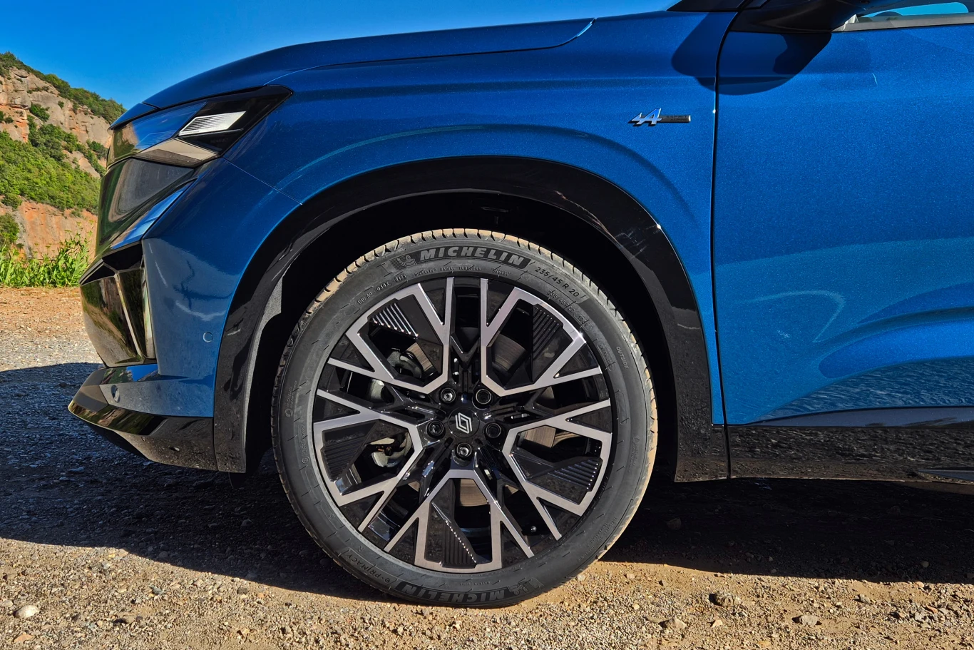 Przednie koło samochodu osobowego z charakterystyczną felgą aluminiową i oponą Michelin, karoseria pojazdu w kolorze niebieskim z widocznym detalem oznaczenia modelu, tło z nieostrymi elementami natury oraz drogą.