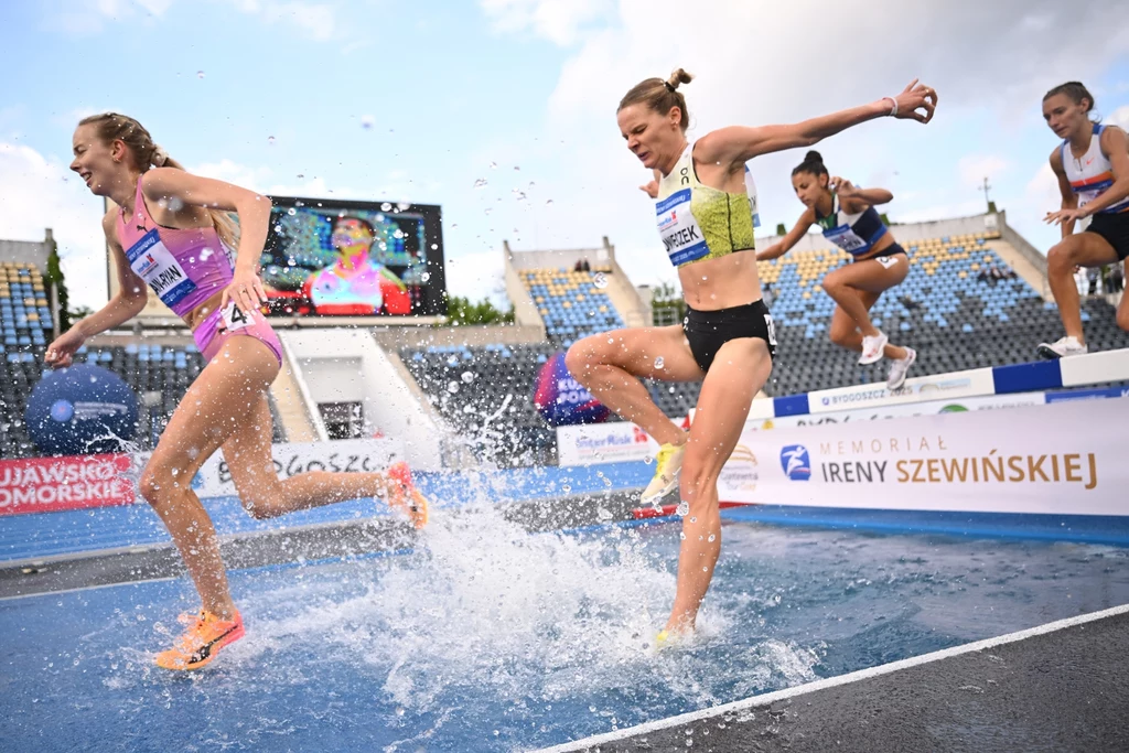 Alicja Konieczek Zawodniczki rywalizujące w biegu z przeszkodami przeskakują przez wodny rów na stadionie lekkoatletycznym, w tle widoczne trybuny i elektroniczny ekran.