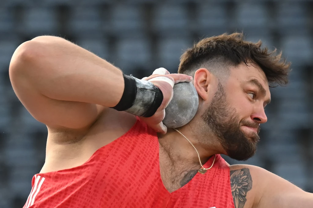 Konrad Bukowiecki w Bydgoszczy Sportowiec w czerwonym stroju wykonuje rzut kulą, koncentrując się na precyzyjnym ruchu, w tle rozmyte trybuny stadionu.
