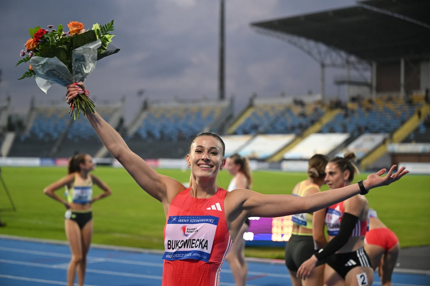 Zawodniczka w czerwonym stroju lekkoatletycznym z bukietem kwiatów unosi ręce w geście triumfu na tle stadionu z widownią; w tle widać inne sportsmenki i trybuny.