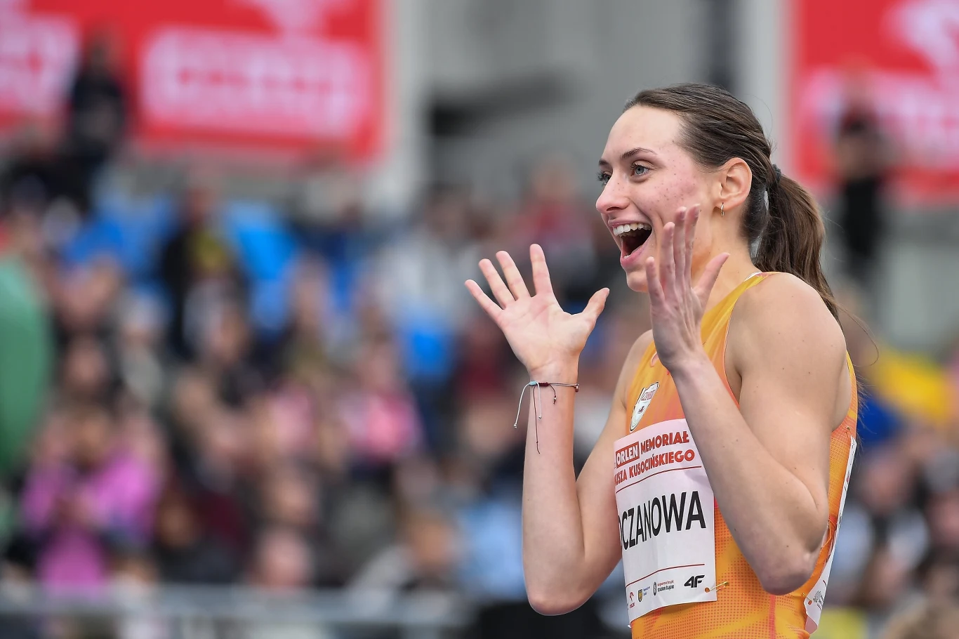 Margarita Koczanowa Uśmiechnięta lekkoatletka w sportowym stroju żółtego koloru z wyraźnym numerem startowym i nazwiskiem, w geście radości unosi otwarte dłonie, w tle niewyraźna publiczność na stadionie i banery reklamowe.