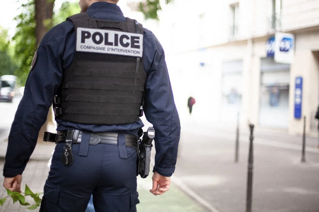Francuska policja udaremniła próbę porwania rodziny biznesmena z branży kryptowalut. Zdj. ilustracyjne Policjant ubrany w kamizelkę kuloodporną z napisem POLICE, stojący tyłem na miejskim chodniku, z widocznym sprzętem policyjnym przy pasku, w tle rozmyte budynki i przechodzień.