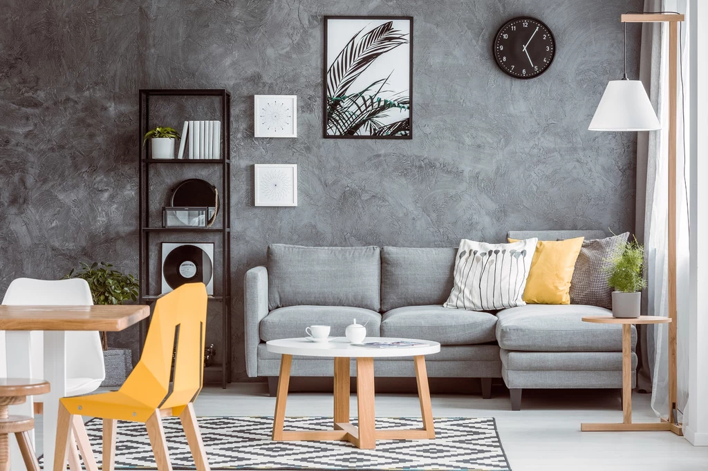 Estetyka millenial grey była popularna w drugiej dekadzie XXI wieku Jasny, nowoczesny salon z szarą sofą, żółtymi akcentami, geometrycznym dywanem, stolikiem kawowym i ozdobnymi obrazami na betonowej ścianie.