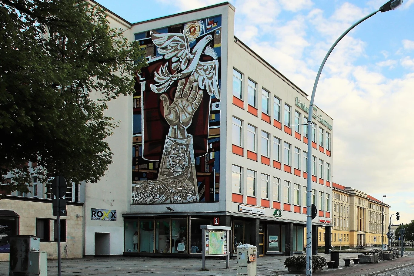 Mozaika ścienna Waltera Womacka na fasadzie dawnego domu towarowego w Eisenhüttenstadt Nowoczesny budynek ozdobiony dużym muralem przedstawiającym wzniesioną dłoń wypuszczającą białego ptaka, symbolizującego wolność lub pokój; poniżej widoczne witryny sklepowe, drzewa i fragment ulicy.