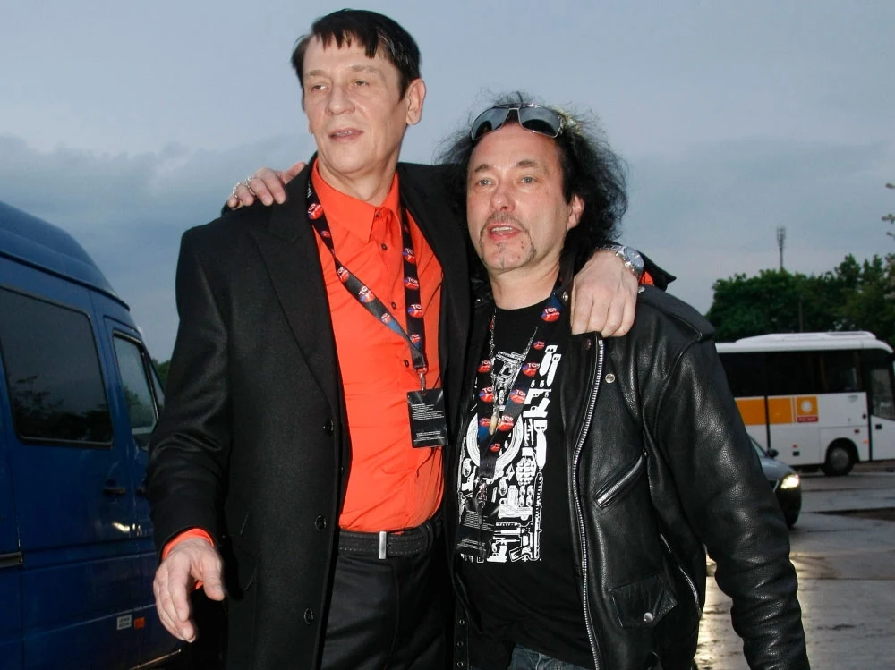 Maciej Maleńczuk i Andrzej Bieniasz (Püdelsi) w 2010 r. Dwóch mężczyzn stoi obok siebie na zewnątrz, jeden ubrany elegancko w czarny garnitur i czerwoną koszulę, drugi ma na sobie skórzaną kurtkę i T-shirt z nadrukiem, obaj mają przypięte identyfikatory na smyczach, w tle zaparkowane samochody i autobus.