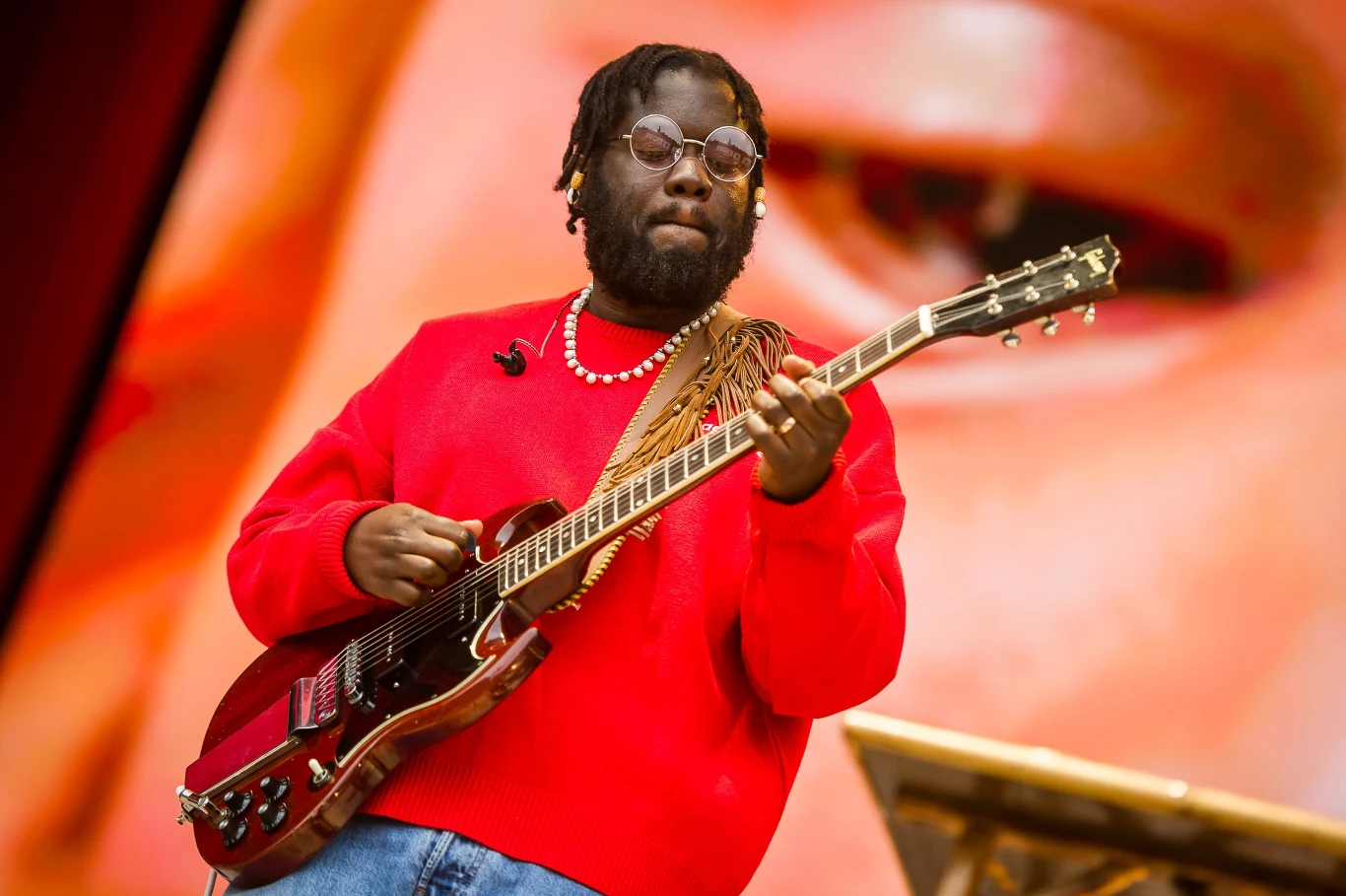 Muzyk w czerwonym swetrze grający na gitarze elektrycznej na tle fragmentu dużego obrazu lub ekranu, wokół stonowane kolory, artysta skupiony na grze, widoczne ozdobne koraliki na szyi.