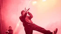 Artysta w ciemnych okularach śpiewający do mikrofonu, wykonujący dynamiczny ruch na scenie oświetlonej intensywnym, czerwonym światłem, z drugą osobą w tle rejestrującą występ telefonem.