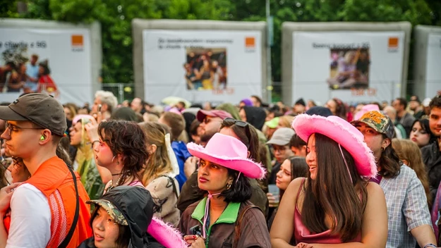 Tłum ludzi zgromadzonych podczas wydarzenia na otwartej przestrzeni; w centrum kobiety w różowych kapeluszach, wokół osoby w ubraniach wierzchnich i festiwalowych akcesoriach, z przodu barierka zabezpieczająca.