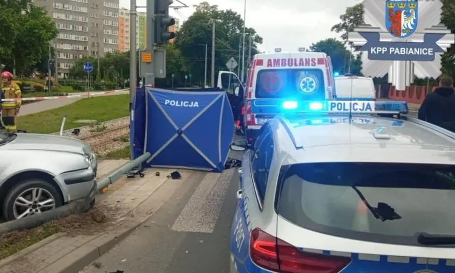 W Pabianicach doszło do tragicznego wypadku Pojazd osobowy uderzony w latarnię uliczną, policyjne auta oraz ambulans stoją przy miejscu zdarzenia, za policyjnym parawanem prowadzone są czynności służb, w tle widoczny budynek mieszkalny i zielone tereny miejskie.