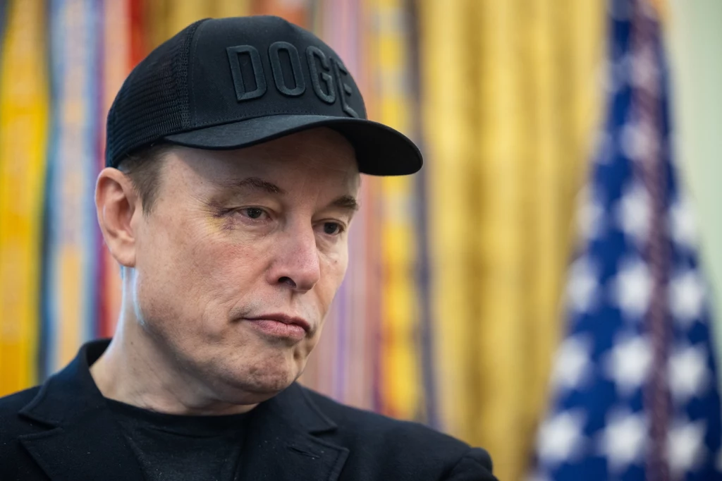 Elon Musk z podbitym okiem przybył na pożegnanie z Białym Domem Mężczyzna w czarnej czapce z napisem DOGE oraz w czarnej marynarce na tle rozmazanej amerykańskiej flagi i żółtych zasłon, wyrażający poważny wyraz twarzy.