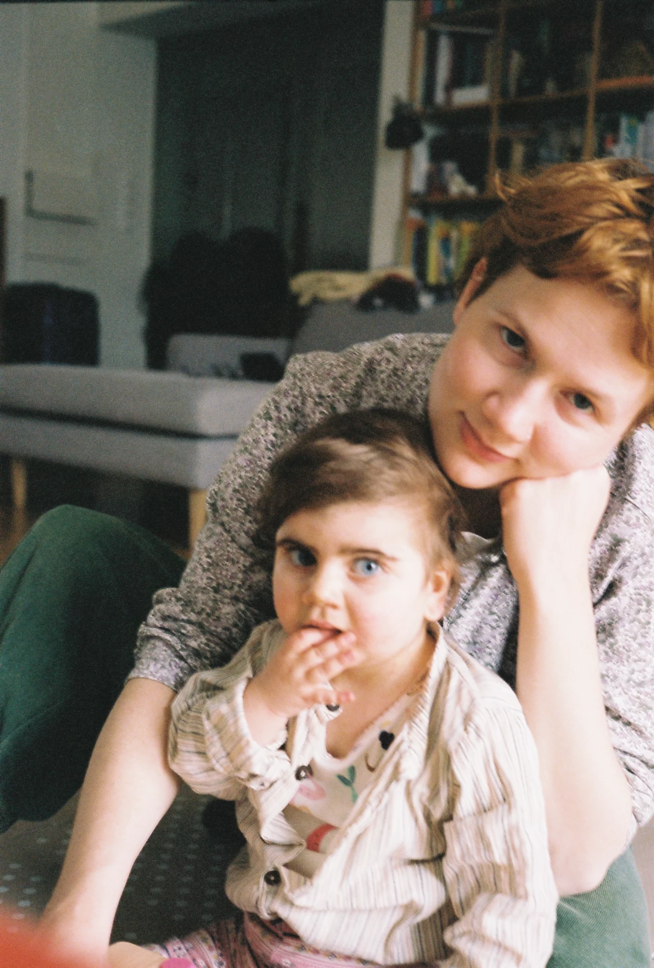 Dorota z córeczką Nadzieją Kobieta z krótkimi rudymi włosami siedząca na podłodze obok dziecka o jasnych oczach i ciemnych włosach, obydwoje zwracają się ku aparatowi, w tle domowe wnętrze z regałem pełnym książek.