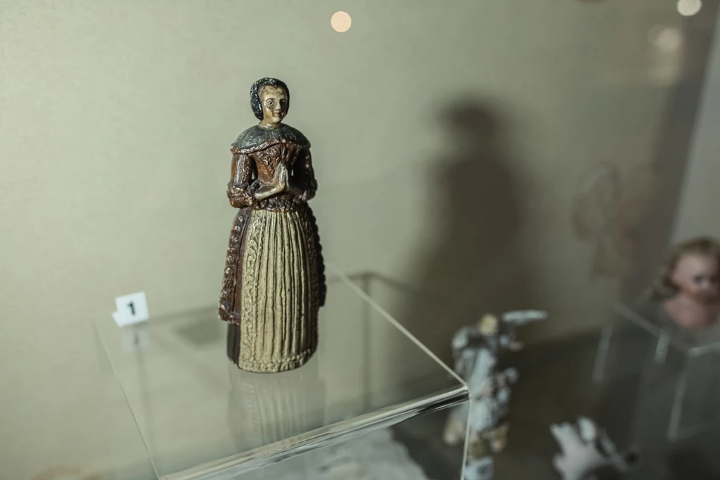 Woskowa lalka, Muzeum Zabawek i Zabawy w Kielcach Stara ceramiczna figurka kobiety w sukni z epoki ustawiona na szklanej gablocie, w tle inne eksponaty i delikatne światło.