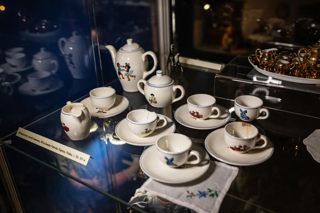 Miniaturowa zastawa, Muzeum Zabawek i Zabawy w Kielcach Zabytkowy zestaw porcelanowych filiżanek i dzbanków do kawy lub herbaty z kolorowymi motywami kwiatowymi ustawiony na półce za szkłem, obok serwetki z haftem, widoczne odbicie światła i dodatkowe złote naczynia w tle.