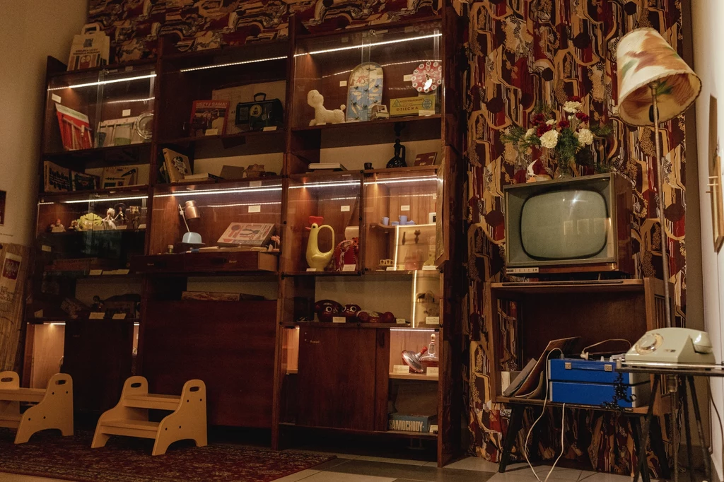 Zbiory Muzeum Zabawek i Zabawy w Kielcach Pokój urządzony w stylu retro z dużą witryną z półkami wypełnionymi zabawkami oraz przedmiotami codziennego użytku z minionej epoki, obok telewizor z lampą i telefonem obrotowym, na tle wzorzystych zasłon.