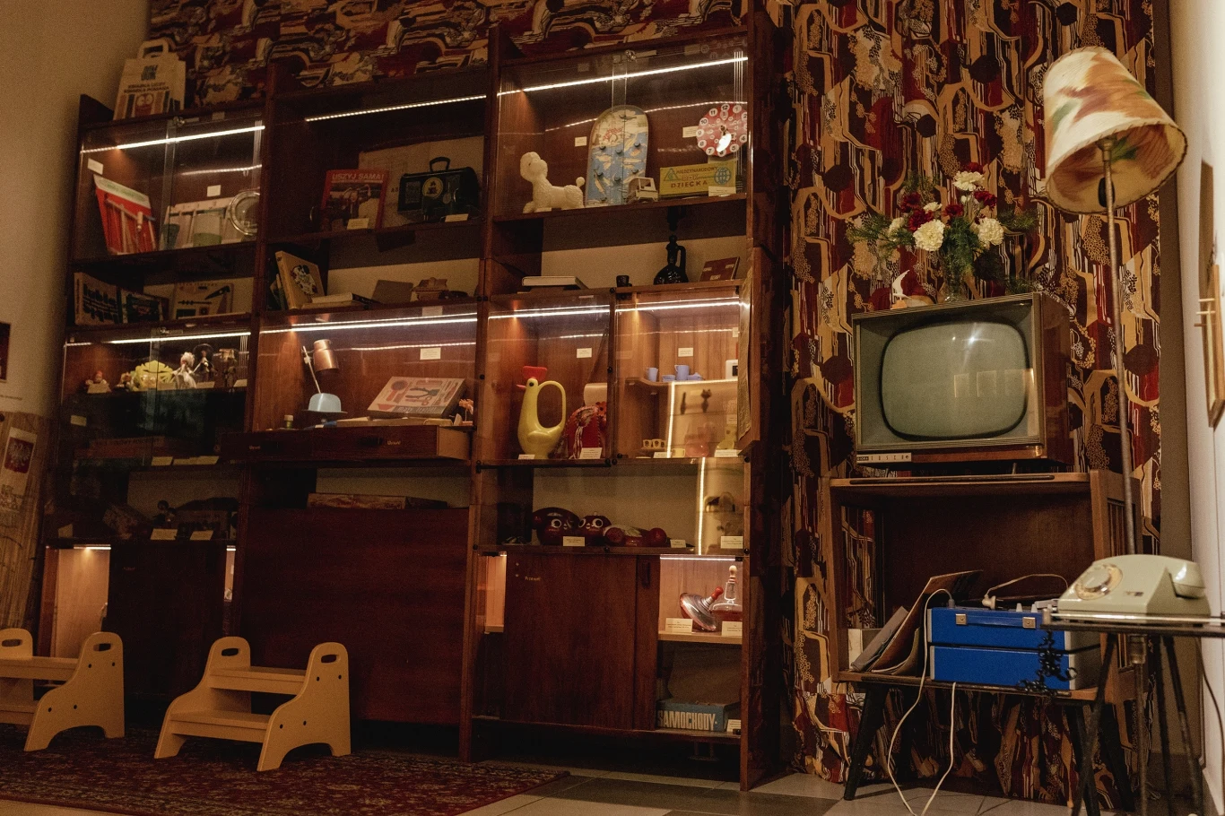 Zbiory Muzeum Zabawek i Zabawy w Kielcach Pokój urządzony w stylu retro z dużą witryną z półkami wypełnionymi zabawkami oraz przedmiotami codziennego użytku z minionej epoki, obok telewizor z lampą i telefonem obrotowym, na tle wzorzystych zasłon.