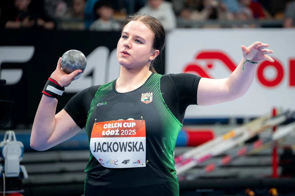 Kinga Jackowska Zawodniczka w trakcie przygotowania do pchnięcia kulą na hali sportowej, ubrana w sportową koszulkę z nazwiskiem i numerem startowym, w tle widoczna część trybun i bannerów reklamowych.