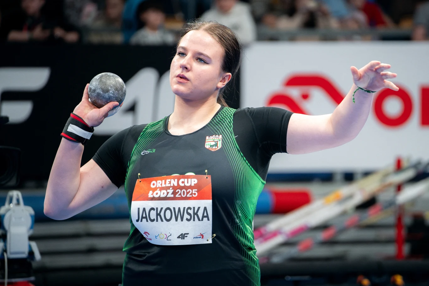 Zawodniczka w trakcie przygotowania do pchnięcia kulą na hali sportowej, ubrana w sportową koszulkę z nazwiskiem i numerem startowym, w tle widoczna część trybun i bannerów reklamowych.