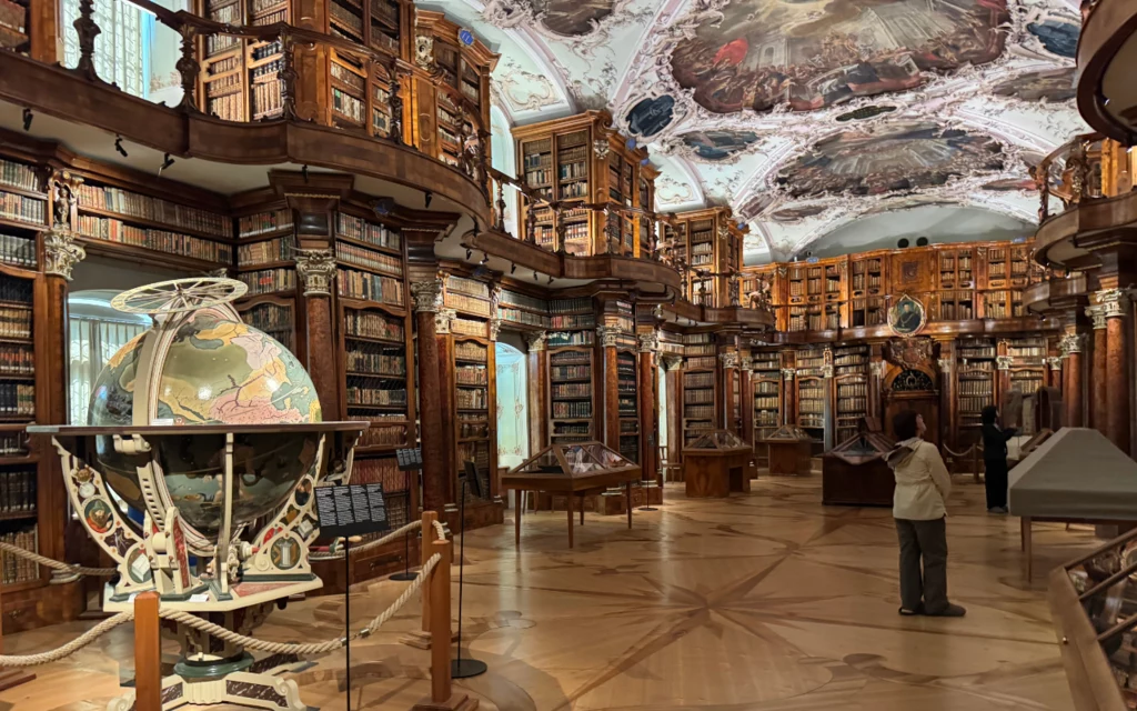 Biblioteka w Opactwie Sankt Gallen Bogato zdobione wnętrze biblioteki z wysokimi regałami pełnymi książek, dużym globusem na pierwszym planie i osobą przyglądającą się eksponatom, klasyczne malowidła na suficie dodają historycznego charakteru.