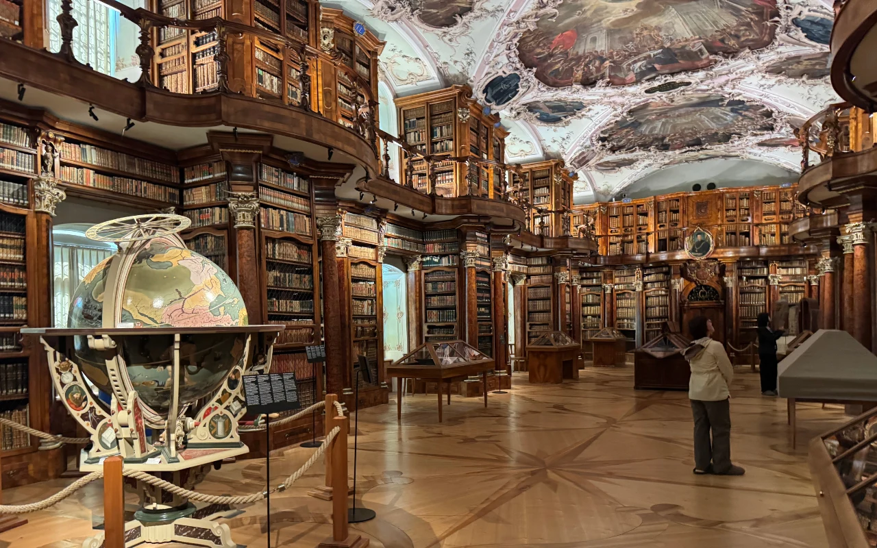 Bogato zdobione wnętrze biblioteki z wysokimi regałami pełnymi książek, dużym globusem na pierwszym planie i osobą przyglądającą się eksponatom, klasyczne malowidła na suficie dodają historycznego charakteru.