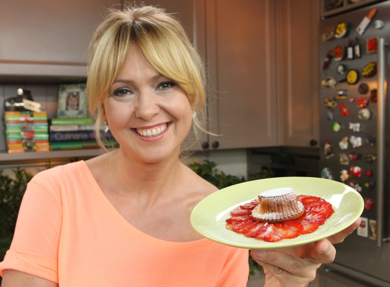 Ewa Wachowicz poleca przysmaki z truskawkami Kobieta z jasnymi włosami w związanych w luźny kok, uśmiechnięta, trzyma talerzyk z deserem z truskawkami i cukrem pudrem, w tle widoczne książki kucharskie oraz lodówka z magnesami.