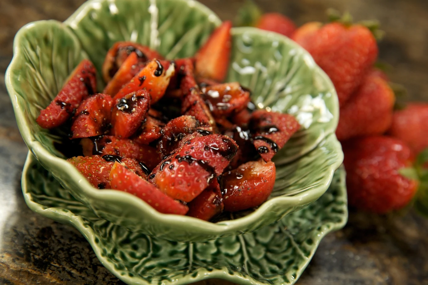 Truskawki z octem balsamicznym Truskawki pokrojone w ćwiartki, polane ciemnym sosem balsamicznym, ułożone w zielonej miseczce ozdobionej wzorem liści, w tle kilka całych truskawek.