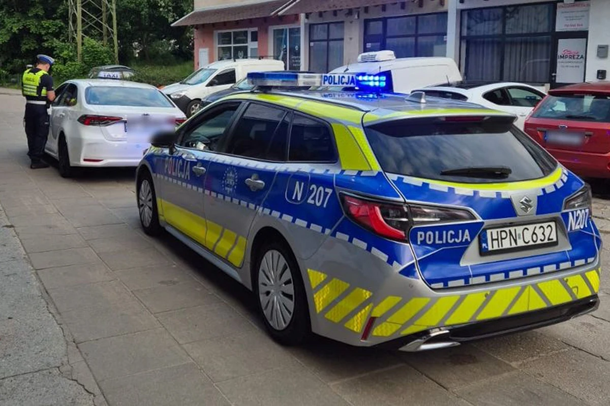 Policjanci przeprowadzili masowe kontrole kierowców wykonujących płatne przewozy "na aplikację" w Trójmieście Radiowóz policyjny z włączonymi światłami stoi zaparkowany za białym samochodem, obok którego policjant w mundurze rozmawia z kierowcą. W tle widoczne są inne samochody oraz budynek z dużymi oknami.