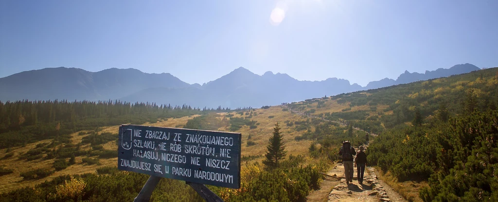 Kiedyś większy był szacunek do gór Dwóch turystów idących górskim szlakiem wśród zielonych łąk i krzewów, w tle masywne pasmo górskie pod błękitnym niebem, z przodu tablica z ostrzeżeniem i zasadami poruszania się po parku narodowym.