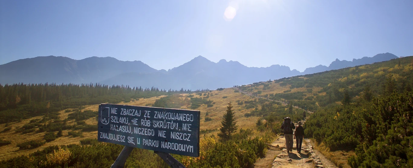Kiedyś większy był szacunek do gór Dwóch turystów idących górskim szlakiem wśród zielonych łąk i krzewów, w tle masywne pasmo górskie pod błękitnym niebem, z przodu tablica z ostrzeżeniem i zasadami poruszania się po parku narodowym.