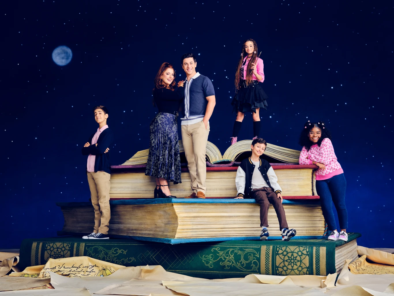 "Czarodzieje z Waverly Place: Nowy rozdział" Grupa sześciu młodych osób pozuje na stosie dużych, otwartych książek, wokół nich rozpościera się nocne niebo z widocznymi gwiazdami i księżycem, osoby ubrane są modnie i radośnie pozują, każda przyjmuje inną pozę podkreślając indywidualny charakter.