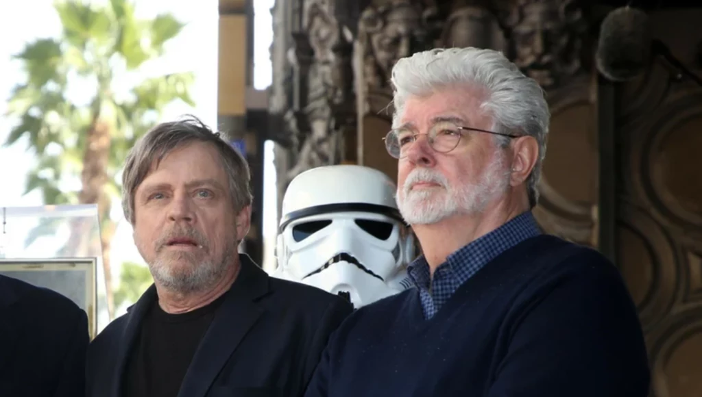 Mark Hamill Dwóch mężczyzn stojących obok siebie, pomiędzy nimi znajduje się postać w kostiumie szturmowca z serii Gwiezdne wojny, w tle widoczna dekoracyjna architektura oraz fragmenty palm.