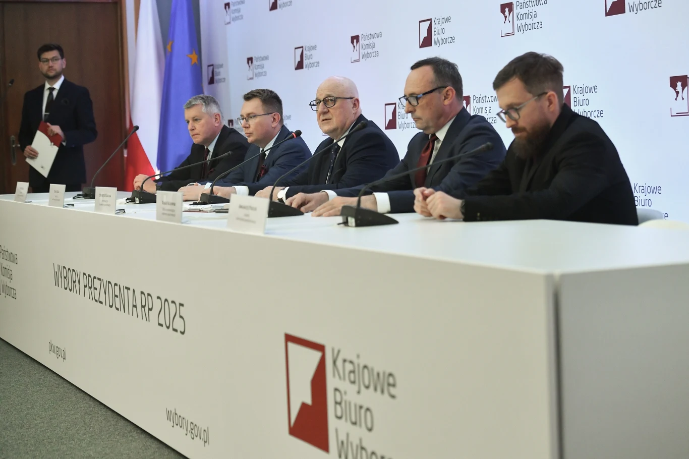 PKW poda dane o frekwencji w wyborach prezydenckich Konferencja prasowa zorganizowana przez Krajowe Biuro Wyborcze, pięciu mężczyzn siedzi za stołem z mikrofonami, na ścianie tło ze znakami instytucji oraz flagi Polski i Unii Europejskiej, obok stoi prowadzący.