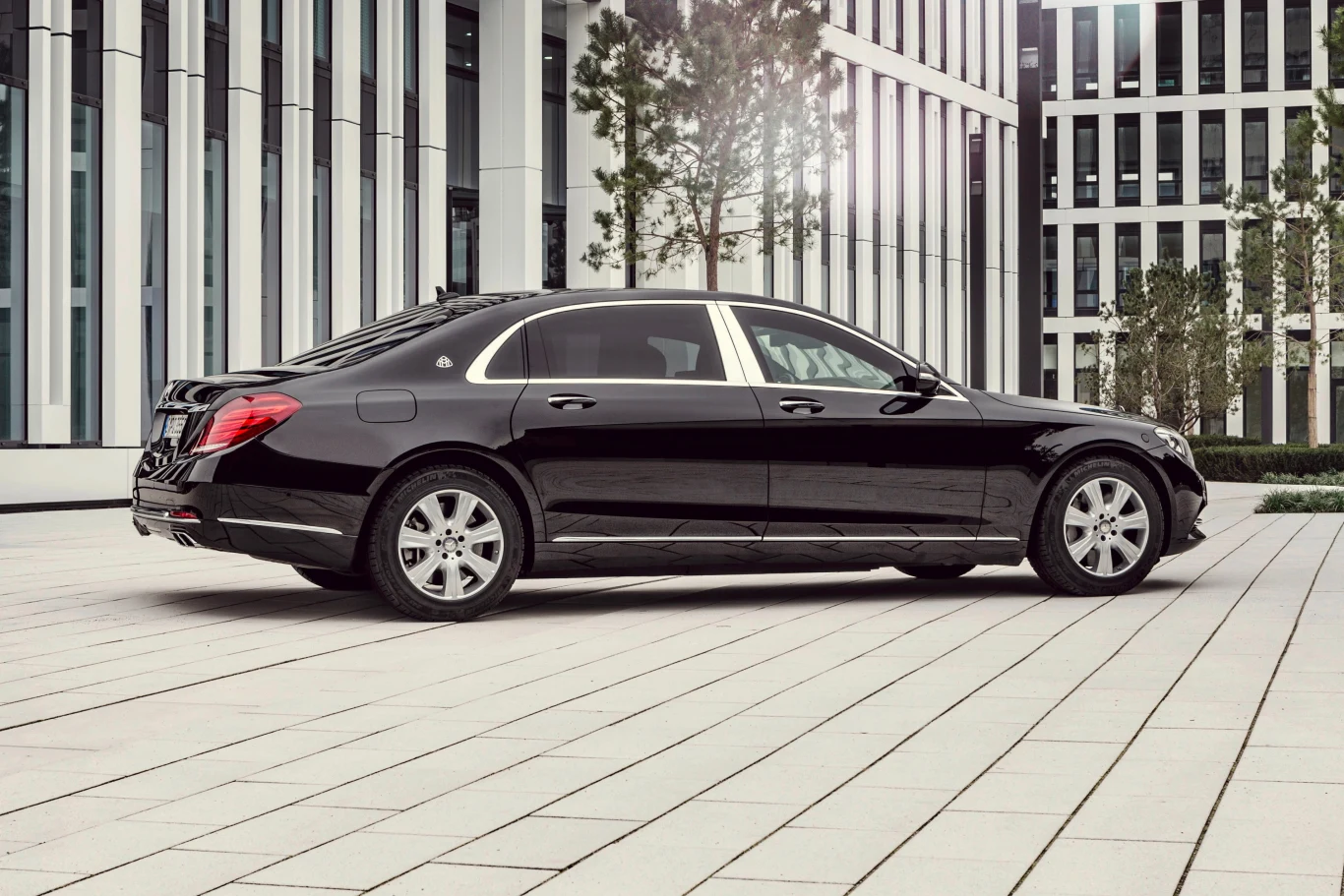 Luksusowa czarna limuzyna, prawdopodobnie marki Mercedes-Maybach, zaparkowana na tle nowoczesnych przeszklonych budynków biurowych oraz zielonych drzew pośród miejskiej architektury.