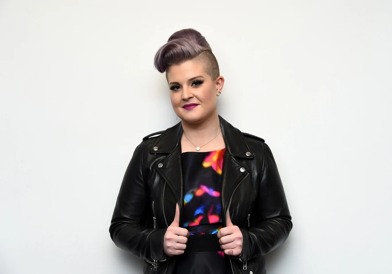 Kelly Osbourne przeszła diametralną metamorfozę - fani jej nie poznają Młoda kobieta z fioletowymi włosami uczesanymi w ekstrawagancką fryzurę z wygolonym bokiem głowy, ubrana w czarną skórzaną kurtkę i kolorową sukienkę, stoi na tle białej ściany, lekko uśmiechnięta, z dłońmi trzymającymi klapy kurtki.