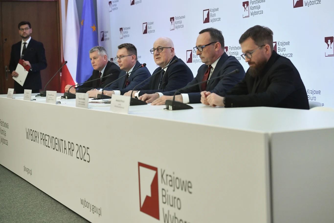 PKW: nie da się zagłosować wielokrotnie poza miejscem zamieszkania Pięciu mężczyzn siedzi za długim stołem konferencyjnym w sali prasowej Krajowego Biura Wyborczego, w tle widoczne flagi Polski i Unii Europejskiej oraz logotypy i napisy dotyczące wyborów prezydenckich 2025 roku.